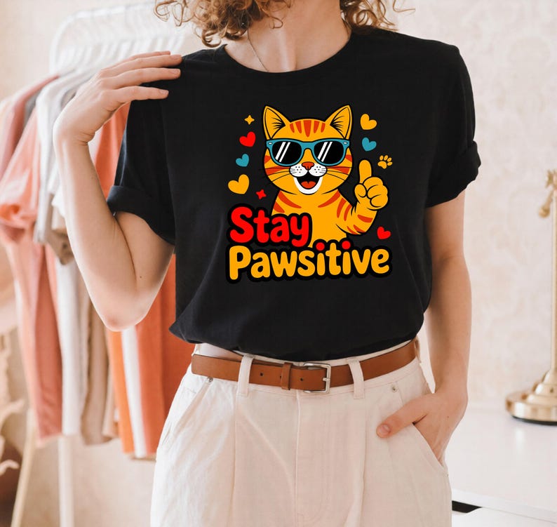 Pode incluir: Camiseta preta com um gato tabby laranja de desenho animado usando &oacute;culos de sol e com o polegar para cima. O texto "Stay Pawsitive" est&aacute; em amarelo e vermelho. O design inclui cora&ccedil;&otilde;es e pegadas. Design divertido.
