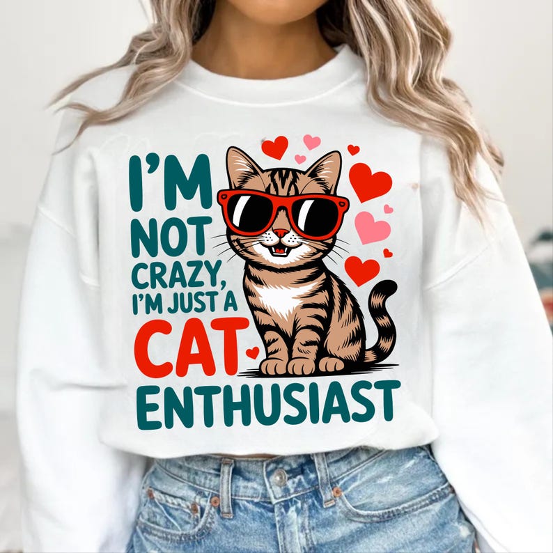 Pode incluir: Camisola branca com um gato de desenho animado usando &oacute;culos de sol vermelhos e cora&ccedil;&otilde;es. O texto diz "I'M NOT CRAZY, I'M JUST A CAT ENTHUSIAST" em verde-azulado e vermelho. Uma pe&ccedil;a confort&aacute;vel para os amantes de gatos.