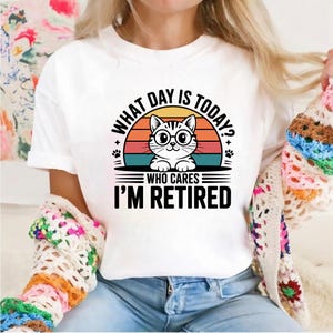 Pode incluir: Camiseta branca com um gr&aacute;fico de um gato usando &oacute;culos, em um design de p&ocirc;r do sol. O texto diz "WHAT DAY IS TODAY? WHO CARES I'M RETIRED". O design inclui pegadas e um esquema de cores retr&ocirc;.