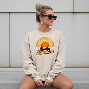 May include: A cream-coloured sweatshirt with a retro sun graphic and the text "Be the Sunshine" in yellow and orange. The sweatshirt has a crew neck and long sleeves. The style is casual and relaxed, suitable for everyday wear.