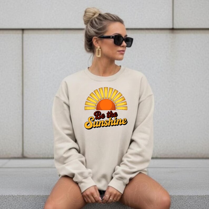 May include: A cream-coloured sweatshirt with a retro sun graphic and the text "Be the Sunshine" in yellow and orange. The sweatshirt has a crew neck and long sleeves. The style is casual and relaxed, suitable for everyday wear.