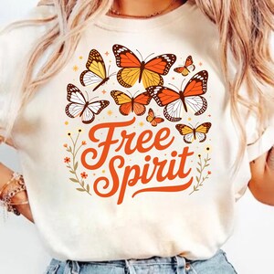 May include: A cream-coloured t-shirt with a design of orange, yellow, and white butterflies. The words "Free Spirit" are written in orange script. The design includes small floral and star accents, creating a whimsical and nature-inspired aesthetic.