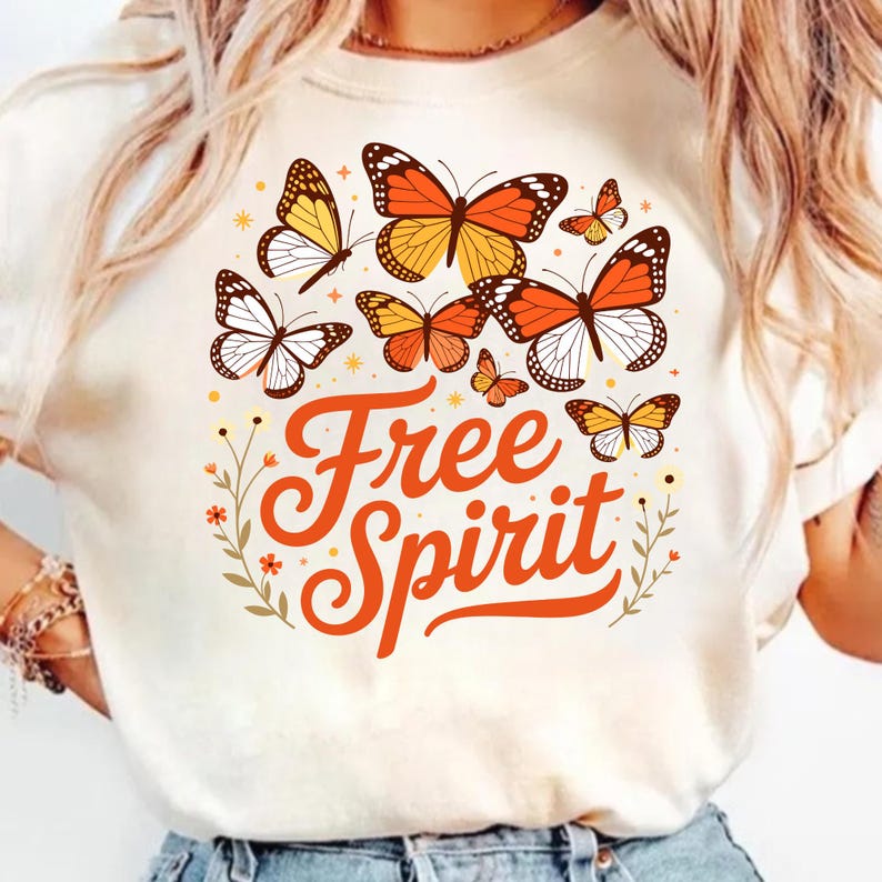 May include: A cream-coloured t-shirt with a design of orange, yellow, and white butterflies. The words "Free Spirit" are written in orange script. The design includes small floral and star accents, creating a whimsical and nature-inspired aesthetic.
