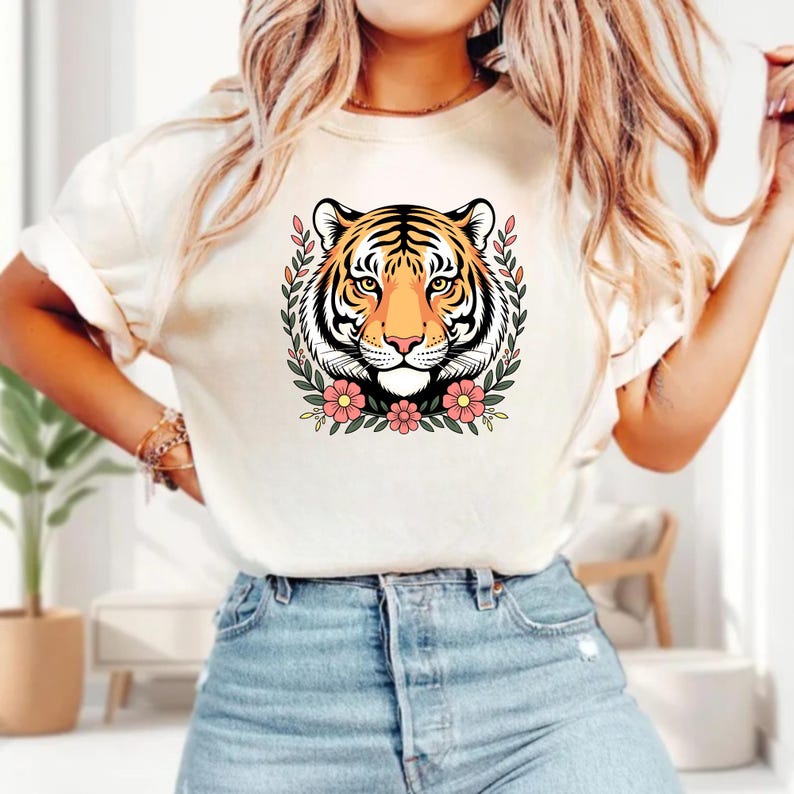 May include: A cream-coloured t-shirt with a tiger's face surrounded by a floral wreath. The tiger has orange, black, and white stripes, with pink flowers and green leaves. The shirt is a casual, everyday wear item.