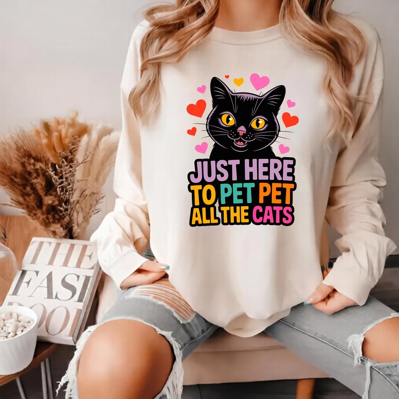 Pode incluir: Camiseta de manga comprida creme com um gr&aacute;fico de gato preto com olhos amarelos e detalhes rosa. O texto "JUST HERE TO PET PET ALL THE CATS" est&aacute; impresso abaixo. Cora&ccedil;&otilde;es vermelhos e rosa circundam o gato.
