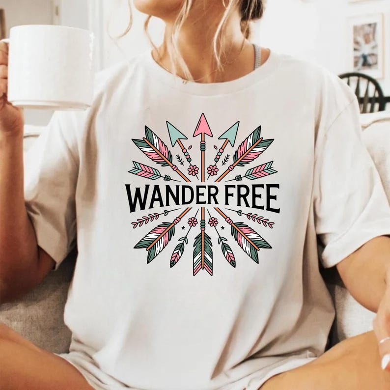 May include: A cream-coloured t-shirt with a circular design of arrows, feathers, and floral elements in pink, teal, and green. The words "WANDER FREE" are printed in black. The shirt is a casual style.