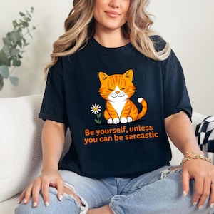 Pode incluir: Camiseta preta com um gato tigrado laranja de desenho animado e uma flor branca. O texto diz: "Seja voc&ecirc; mesmo, a menos que possa ser sarc&aacute;stico". A camiseta &eacute; usada por uma pessoa.