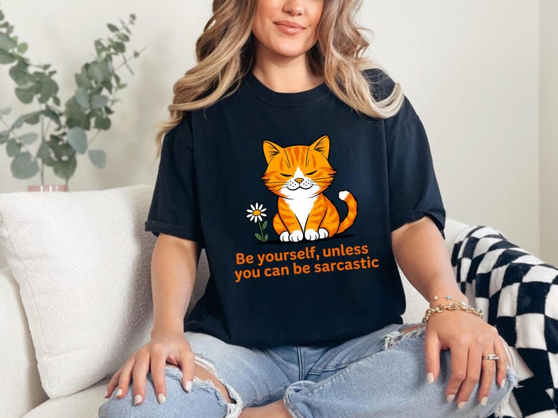 Pode incluir: Camiseta preta com um gato tigrado laranja de desenho animado e uma flor branca. O texto diz: "Seja voc&ecirc; mesmo, a menos que possa ser sarc&aacute;stico". A camiseta &eacute; usada por uma pessoa.