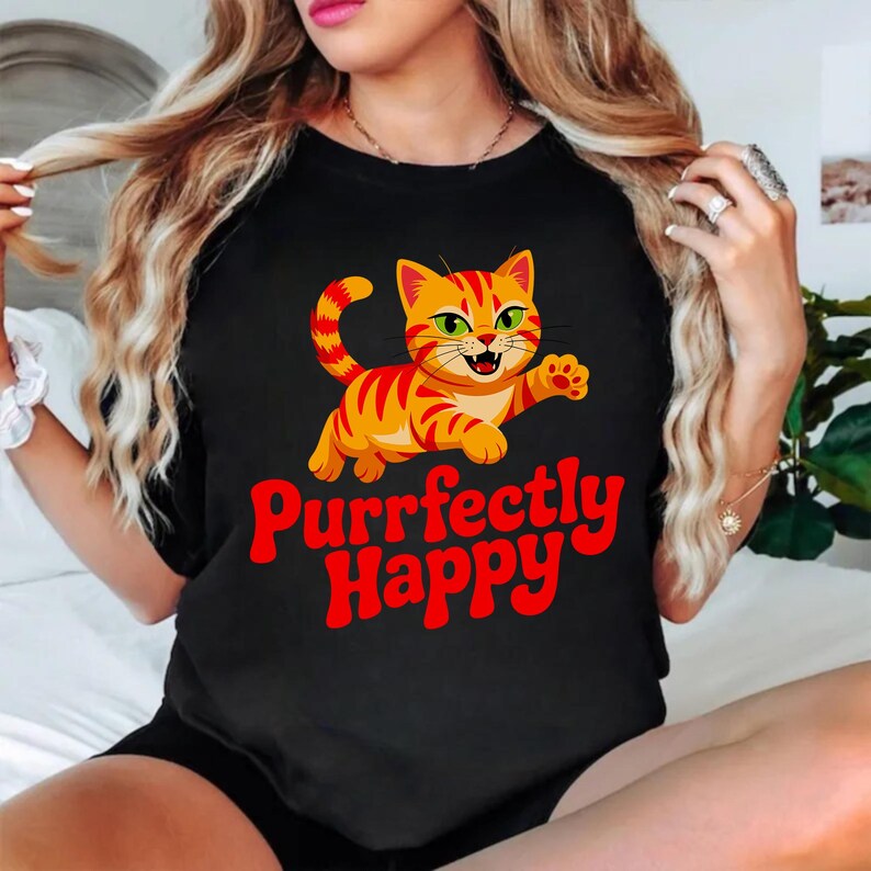 Pode incluir: Camiseta preta com um desenho de gato tigrado laranja de desenho animado com listras vermelhas e olhos verdes. O texto "Purrfectly Happy" est&aacute; em vermelho. &Eacute; uma roupa casual.