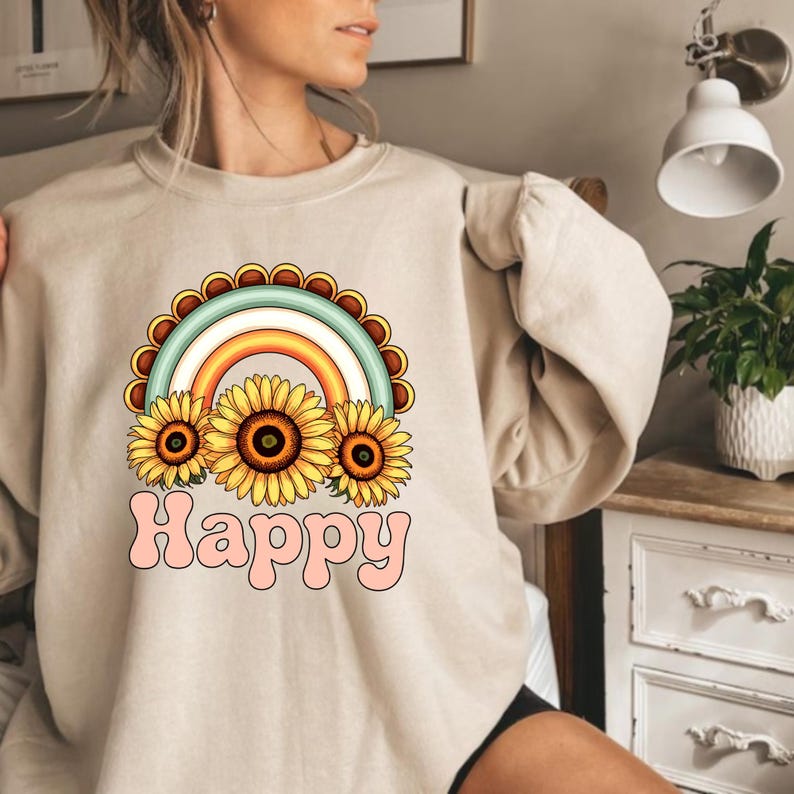 May include: Beige sweatshirt with a colourful graphic of a rainbow, sunflowers, and the word "Happy" in a bubble font. The design includes shades of yellow, orange, blue, and pink. The sweatshirt has long sleeves.