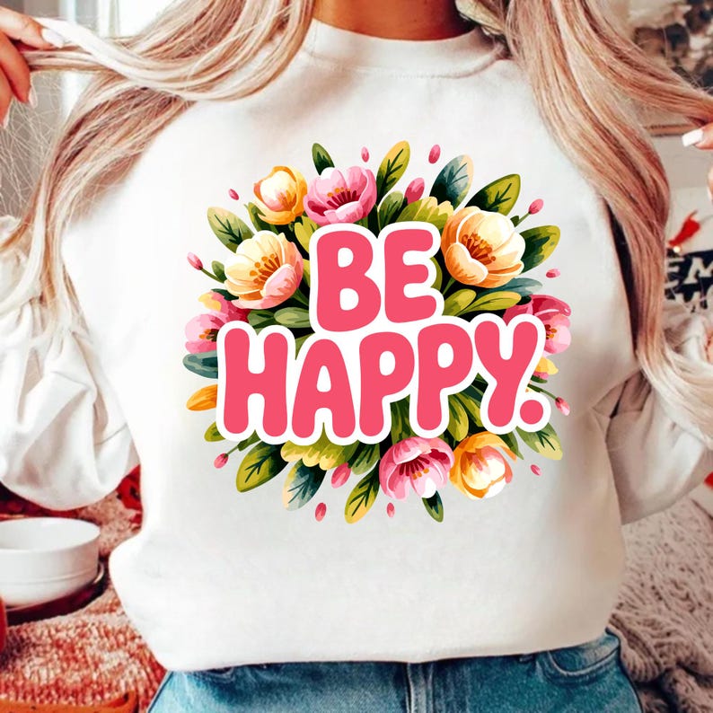 May include: White sweatshirt featuring a floral design and the words "BE HAPPY" in pink. The floral arrangement includes pink, yellow, and orange flowers with green leaves. A casual and cheerful design.