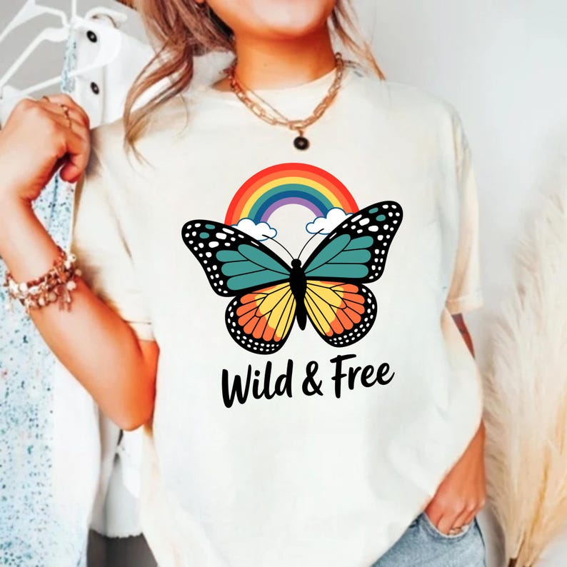 May include: A cream-coloured t-shirt with a colourful butterfly design, rainbow, and the text "Wild & Free". The butterfly has teal, yellow, and orange wings with black detailing. The shirt is a casual style.