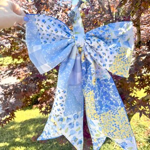 Könnte beinhalten: Eine große dekorative Schleife aus einem Patchwork aus Stoffquadraten mit verschiedenen blauen, weißen und gelben Blumen-, Vichy- und Unimustern. Die Schleife hat zwei lange, spitz zulaufende Enden und einen zentralen Stoffknoten.