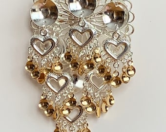 May include: A silver and gold-toned brooch with a filigree design. The brooch features heart-shaped pendants and dangling gold-coloured beads. The top has three silver discs and a decorative openwork design.