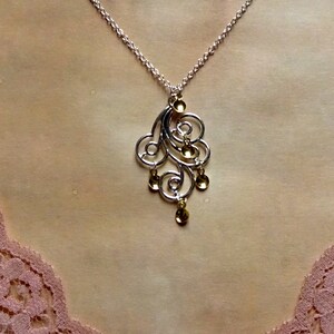 Peut inclure: Un collier en argent avec un pendentif en or pr&eacute;sentant un motif d&eacute;licat et tourbillonnant. Le pendentif comporte plusieurs petites accents ronds en or.