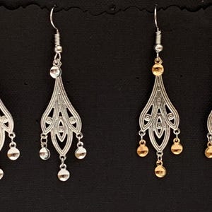 Puede incluir: Un par de pendientes colgantes con acabado plateado y dorado. Los pendientes presentan un diseño delicado e intrincado con múltiples niveles de pequeñas cuentas redondas.