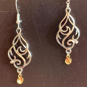 Puede incluir: Un par de pendientes de plata con una cuenta dorada en la parte inferior. Los pendientes tienen un diseño delicado e intrincado.