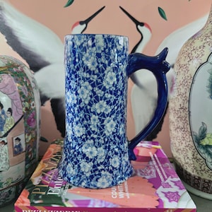 Può includere: Un boccale da birra a fantasia floreale blu e bianca con un manico blu scuro. Il boccale è appoggiato su una pila di libri, tra cui "PIZZAZZERIE Entertain in Style" e "GARDENTOPIA". Lo sfondo presenta una parete rosa con un disegno di gru.