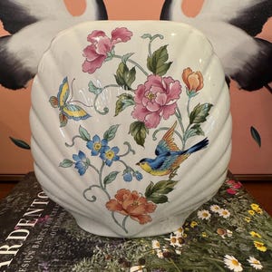 May include: Cream-colored, shell-shaped vase with floral and bird design. The vase features pink peonies, blue and orange flowers, green leaves, a blue bird, and a yellow and blue butterfly. The vase is sitting on a book.