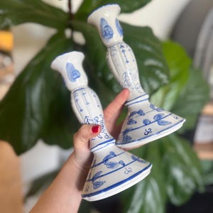 May include: Two white ceramic candlesticks with blue floral patterns. The candlesticks have a flared base, a long stem, and a flared top. The blue floral design includes leaves and other decorative elements. The candlesticks are held in front of a green leafy background.