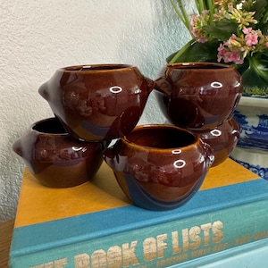 Puede incluir: Un conjunto de cinco cuencos pequeños de cerámica marrón brillante con asas. Los cuencos están colocados encima de un libro con letras doradas. Los cuencos tienen un diseño clásico y vintage, probablemente para servir sopa.