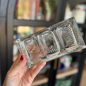 May include: A clear, rectangular glass serving dish with three compartments. The dish has a textured, fan-like pattern in each compartment. The edges are slightly raised. The dish is held up in front of a blurred background.