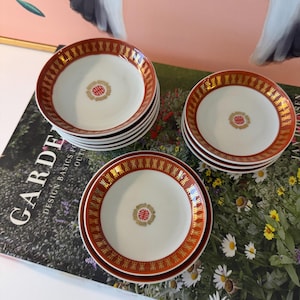 May include: A collection of small, white ceramic plates with a red and gold rim design. Each plate features a gold emblem in the center. The plates are stacked in groups, showcasing a classic, elegant aesthetic.