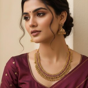 May include: A gold-toned necklace featuring a row of small, oval-shaped magenta stones set in intricate metalwork with dangling gold beads, paired with matching bell-shaped jhumka earrings that have a textured gold finish and small hanging spheres.