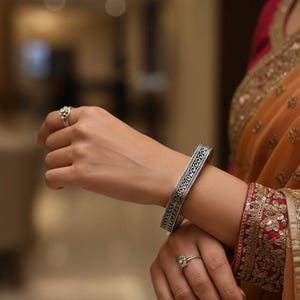 May include: A silver-toned metal bangle bracelet featuring an intricate embossed pattern along its surface, worn alongside two silver-toned rings with small clear stones, paired with an orange and magenta fabric garment with gold embroidery.