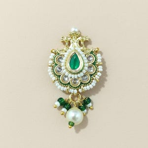 May include: A gold-tone brooch featuring a central teardrop-shaped green stone surrounded by clear crystals and white seed beads. The design includes intricate metalwork, green accent beads, and a single large white pearl-like bead dangling below.