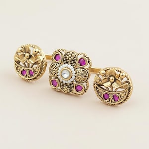 May include: Three gold-colored rings with floral and geometric designs. The rings feature small, round, and square shapes with pink gemstones and a white center stone. The rings are set against a light background.