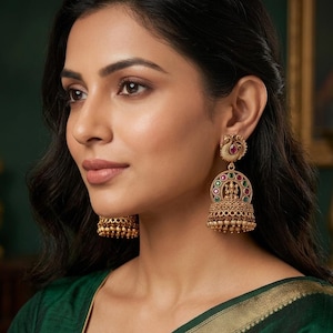 May include: Gold-toned jhumka earrings featuring a peacock-shaped stud with a central red stone. The bell-shaped drop displays a central deity figure surrounded by alternating circular red and green stones, finished with small dangling gold beads.