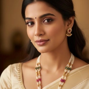 May include: A woman wearing a cream-colored sari and gold jewelry. The jewelry includes a multi-strand pearl necklace with ruby and emerald accents, and matching earrings. The woman has dark hair and a bindi.