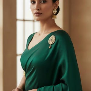 May include: A deep emerald green saree featuring a smooth, matte fabric texture. The garment is adorned with a gold-toned brooch featuring white pearls and a small red accent stone. Matching gold-toned jhumka earrings with pearls are worn.