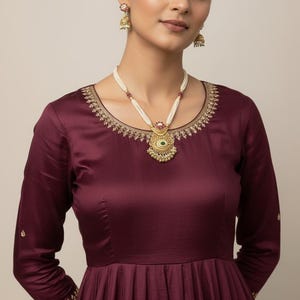 May include: A woman wearing a burgundy dress with long sleeves and gold embroidery. She is adorned with a pearl necklace with a gold pendant and matching earrings. The dress has a pleated skirt.