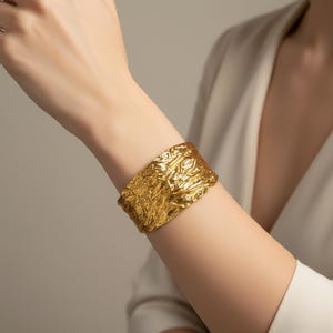 May include: A wide gold cuff bracelet featuring a highly textured, irregular, and organic surface that mimics the appearance of crumpled metal. The bracelet has a polished, reflective finish that catches the light across its uneven ridges.