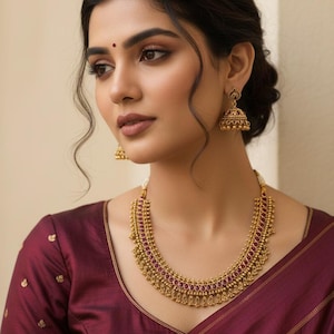 May include: A woman wearing a gold-colored necklace and matching earrings with red gemstone accents. The necklace and earrings are ornate and detailed. She is wearing a maroon-colored blouse.