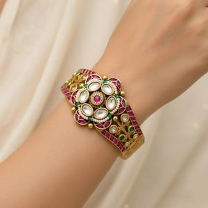 May include: A gold-toned cuff bracelet featuring a central floral motif set with white teardrop stones, a round pink center stone, and small pearls. The band is accented with rectangular pink stones, green accents, and intricate metal filigree detailing.