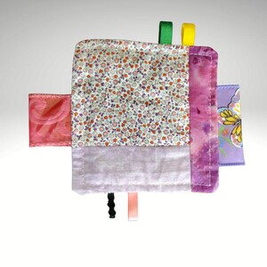 May include: A handmade baby sensory toy with a floral print, purple tie-dye, and lavender fabric. It features colorful ribbons and a patterned purple square. The toy is designed to stimulate a baby's senses.