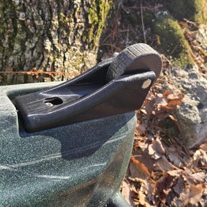 May include: Close-up of a black kayak wheel assembly. The wheel is round and made of a dark material, attached to a black bracket. The kayak's hull is dark green with white scratches.