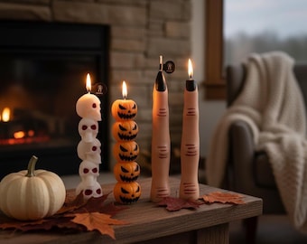 May include: A Halloween-themed display featuring lit candles shaped like skulls, jack-o'-lanterns, and fingers. A small white pumpkin and fall leaves are on a wooden table. The candles are orange, white, and flesh-toned, creating a spooky ambiance.