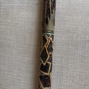 May include: A wooden walking stick featuring a dark brown, glossy finish with a carved, textured pattern. The design includes a section with geometric, diamond-shaped carvings and a smooth, bark-like upper portion with deep, vertical grooves.