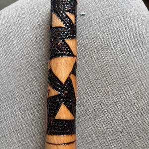 May include: A cylindrical wooden rod featuring a smooth, light brown natural wood finish at the top and bottom ends. The central section displays a contrasting dark, textured, burnt-wood pattern with geometric triangular shapes carved into the surface.