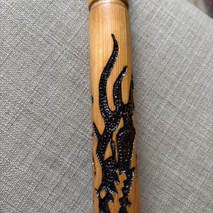 May include: A wooden walking stick with a light brown finish and a dark brown, flame-like design carved into the wood. The top of the stick has a darker, textured pattern.