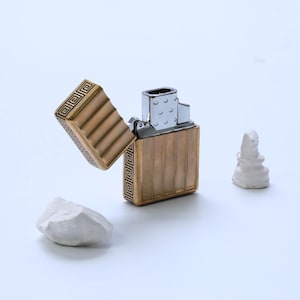 Może przedstawiać: Złoty zapalniczka z prostokątnym korpusem i otwieraną pokrywą. Zapalniczka ma pionowy wzór w paski i dekoracyjne geometryczne wzory. Otwarta pokrywa odsłania wnętrze zapalniczki na białym tle.