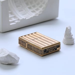 Puede incluir: Un encendedor rectangular de color dorado con una parte superior texturizada y un diseño de llave griega. El encendedor tiene la palabra "SENECA" y la frase "Ignis aurum probat miseria fortes viros" grabadas. También está grabado "Hand Made".