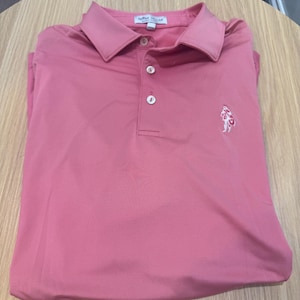 以下が含まれることがあります： 襟付きで白いボタンが3つ付いたピンク色のポロシャツ。胸には小さな白いロゴが付いています。シャツは明るい茶色の木製の表面にきちんと折りたたまれています。