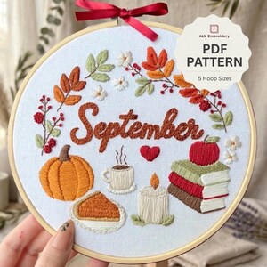 May include: A round wooden embroidery hoop containing white fabric with hand-stitched autumn motifs. The design features the word September in brown, surrounded by a pumpkin, a slice of pie, a steaming mug, a candle, and a stack of books.