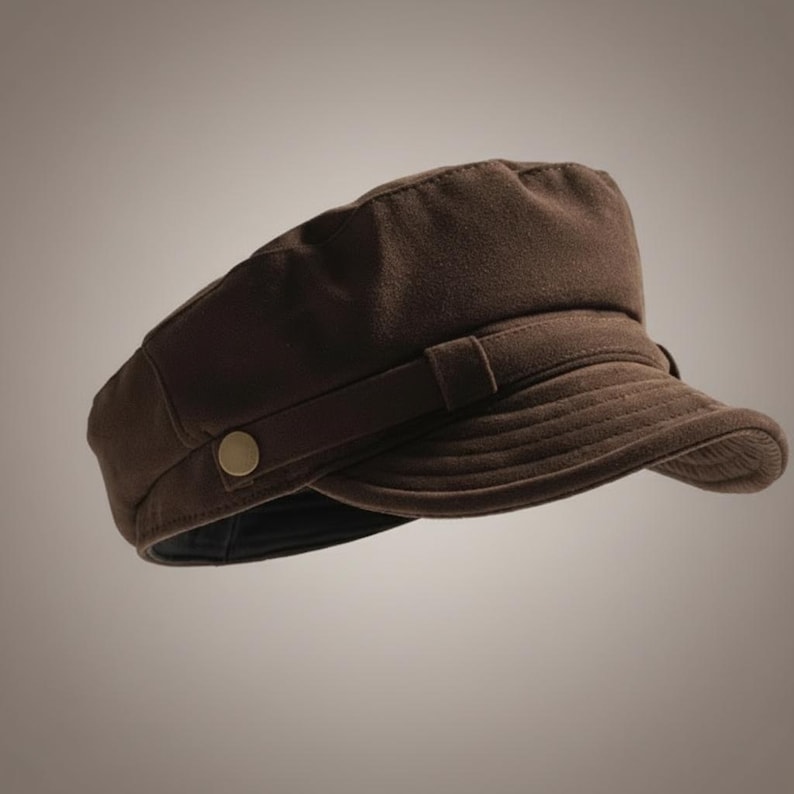 Puede incluir: Gorra marr&oacute;n de estilo newsboy con una corona redondeada y una visera corta y curvada. La gorra presenta un detalle de banda con un bot&oacute;n dorado en el lateral. El sombrero est&aacute; hecho de un material suave y texturizado.