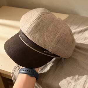 May include: A newsboy cap featuring a light beige, textured fabric crown with subtle vertical stripes and a dark brown, smooth-textured brim. A dark brown band separates the crown and brim, accented by a small, rectangular metallic plate.